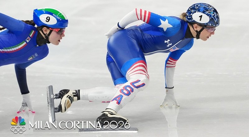 U.S. short track speed skater (number 1) leans low in intense pursuit during a women's race at the Milano Cortina 2026 Winter Olympics, closely trailed by Italy's skater (number 9) in blue suit on the ice rink. NBC Olympics broadcast capture of high-stakes competition.